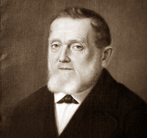 Porträt eines älteren Mannes mit Bart und dunklem Anzug vor neutralem Hintergrund.