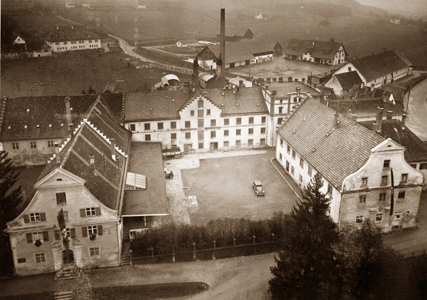 Ein historisches Luftbild zeigt ein Dorf mit verschiedenen Gebäuden und einer Dorfstraße. Ein einzelner Pkw steht auf dem zentralen Platz.