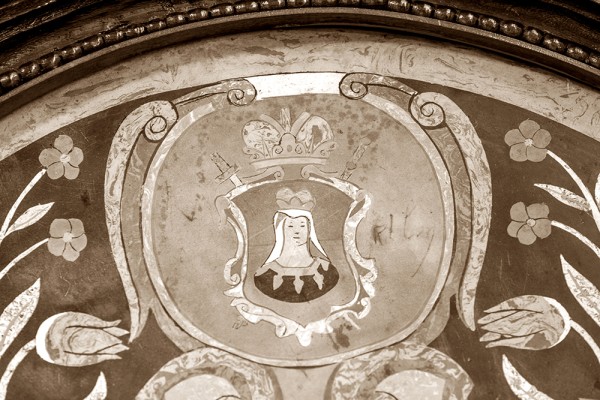 Ein historisches Wappen mit Blumen und einer Maus im Zentrum ist zu sehen, umgeben von dekorativen Elementen.