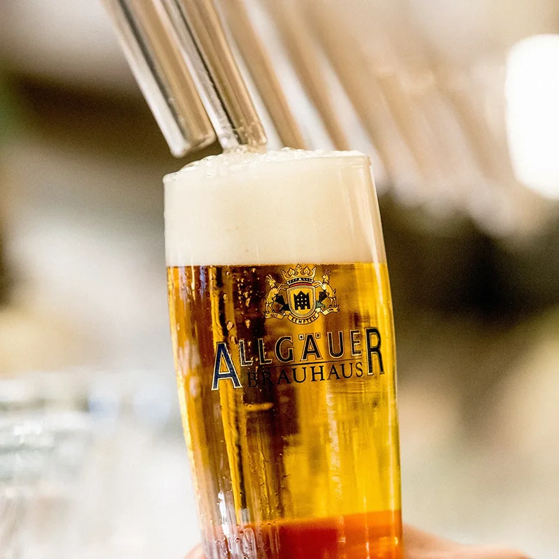 Eine Hand hält ein volles Bierglas mit Schaumkrone, im Hintergrund sind unscharfe Gläser.