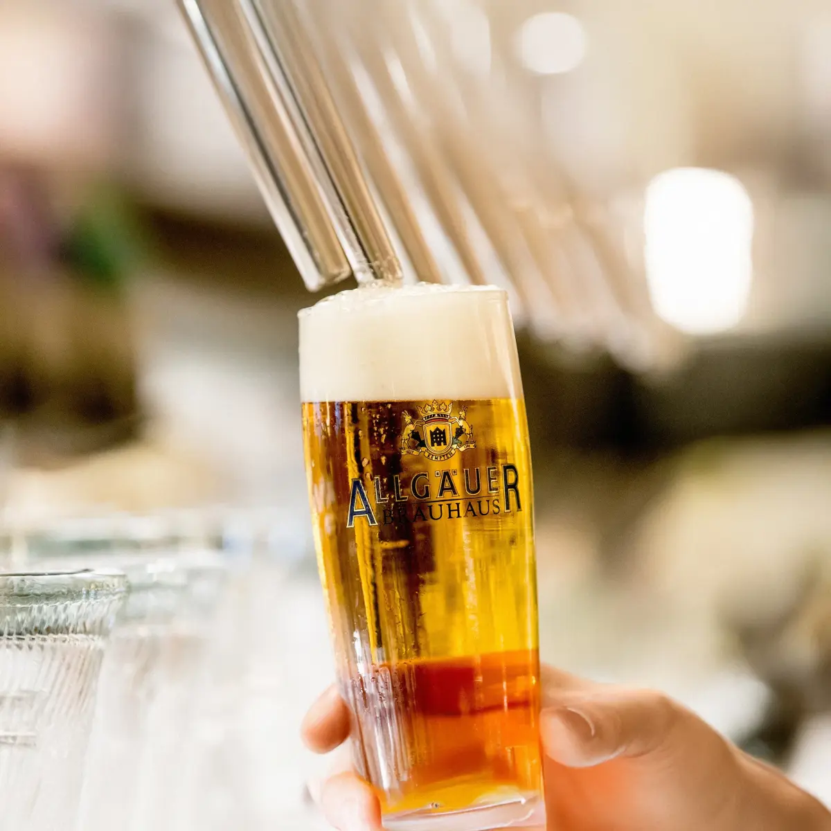Eine Hand hält ein volles Bierglas mit Schaumkrone, im Hintergrund sind unscharfe Gläser.