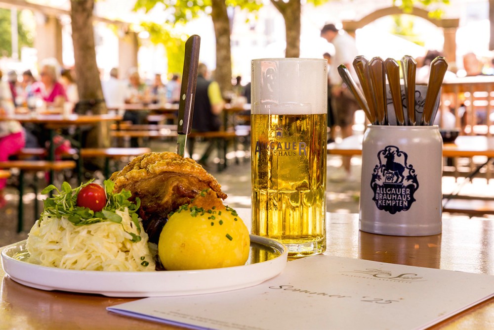 Großaufnahme einer Haxe neben einem Allgäuer Brauhaus Bierglas, im Hintergrund Biergatenatmosphäre.