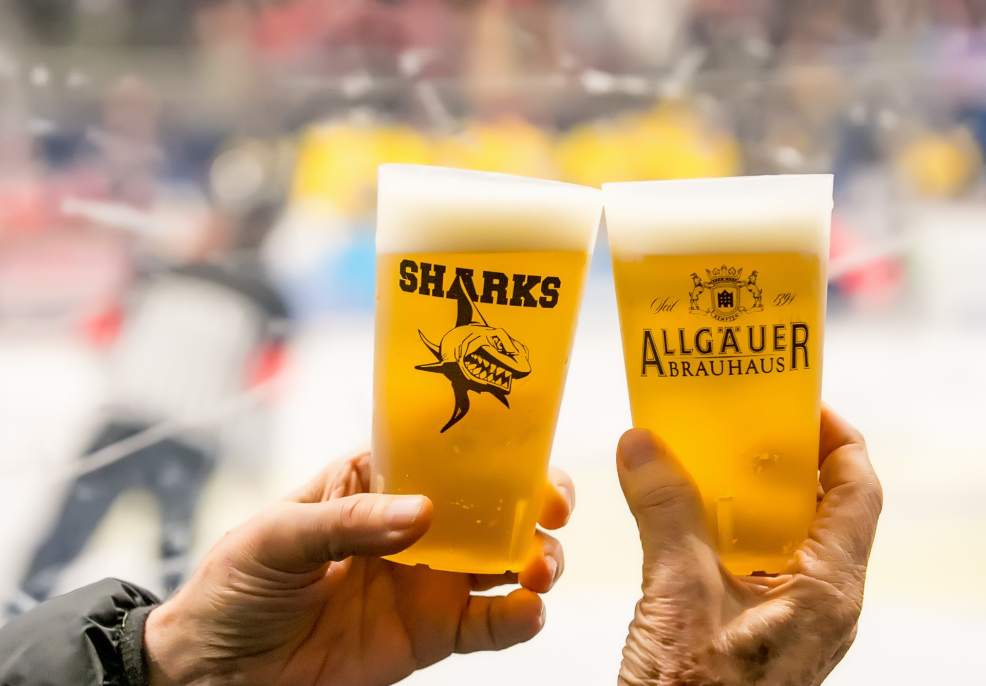 Zwei Hände halten Biergläser mit Logos, im Hintergrund unscharfe Menschen.