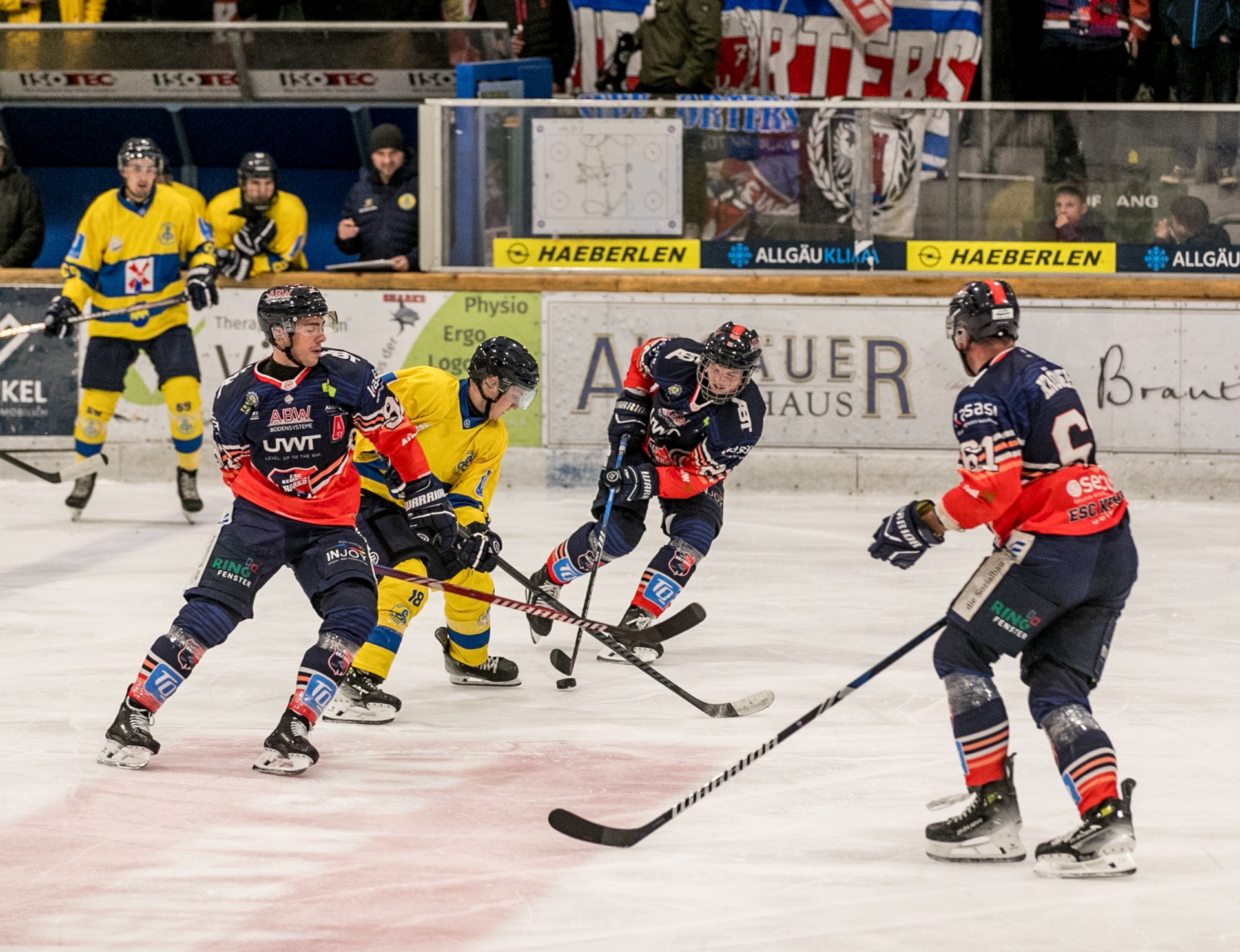 Drei Eishockeyspieler kämpfen um einen Puck auf einer Eisbahn. Zwei Spieler tragen gelbe, einer rote Kleidung.