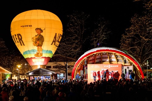 Ein beleuchteter Heißluftballon schwebt über einer Menschenmenge bei einer nächtlichen Veranstaltung.