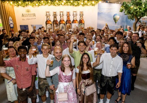 Viele Menschen in traditioneller und moderner Kleidung stoßen mit Biergläsern an. Sie lächeln und feiern gemeinsam in einer Festsituation.