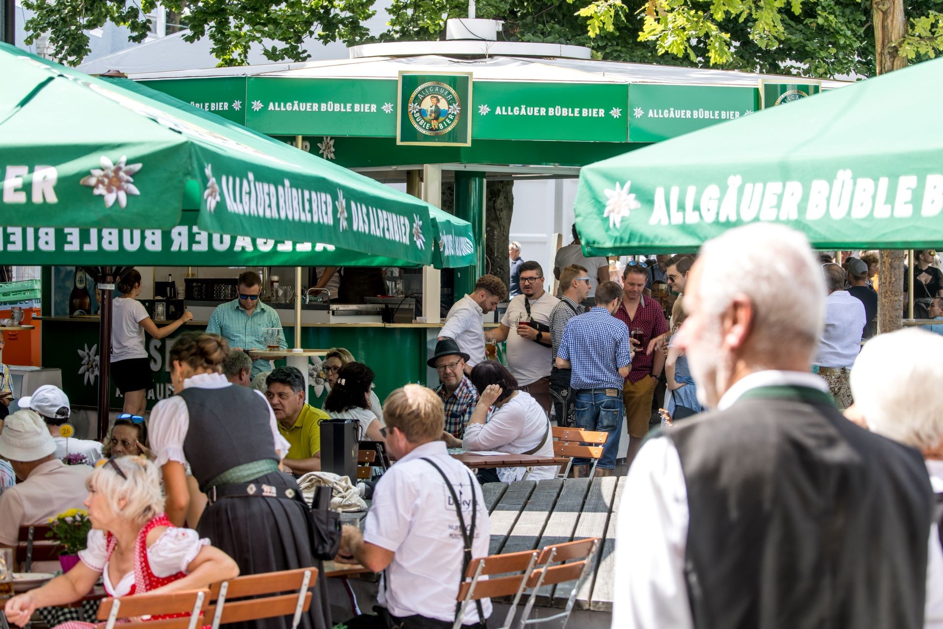 Viele Menschen genießen Getränke und Gespräche an Bierständen unter grünen Sonnenschirmen.