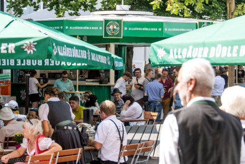 Viele Menschen genießen Getränke und Gespräche an Bierständen unter grünen Sonnenschirmen.