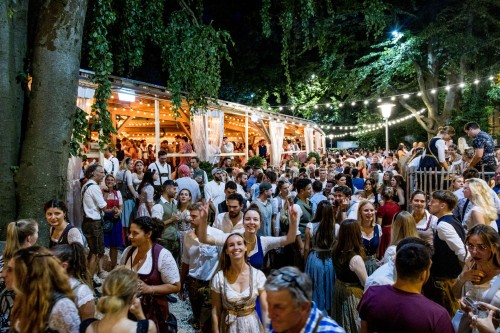 Eine Menschenmenge feiert fröhlich bei einem beleuchteten Abendevent im Freien. Einige tragen traditionelle Trachten.