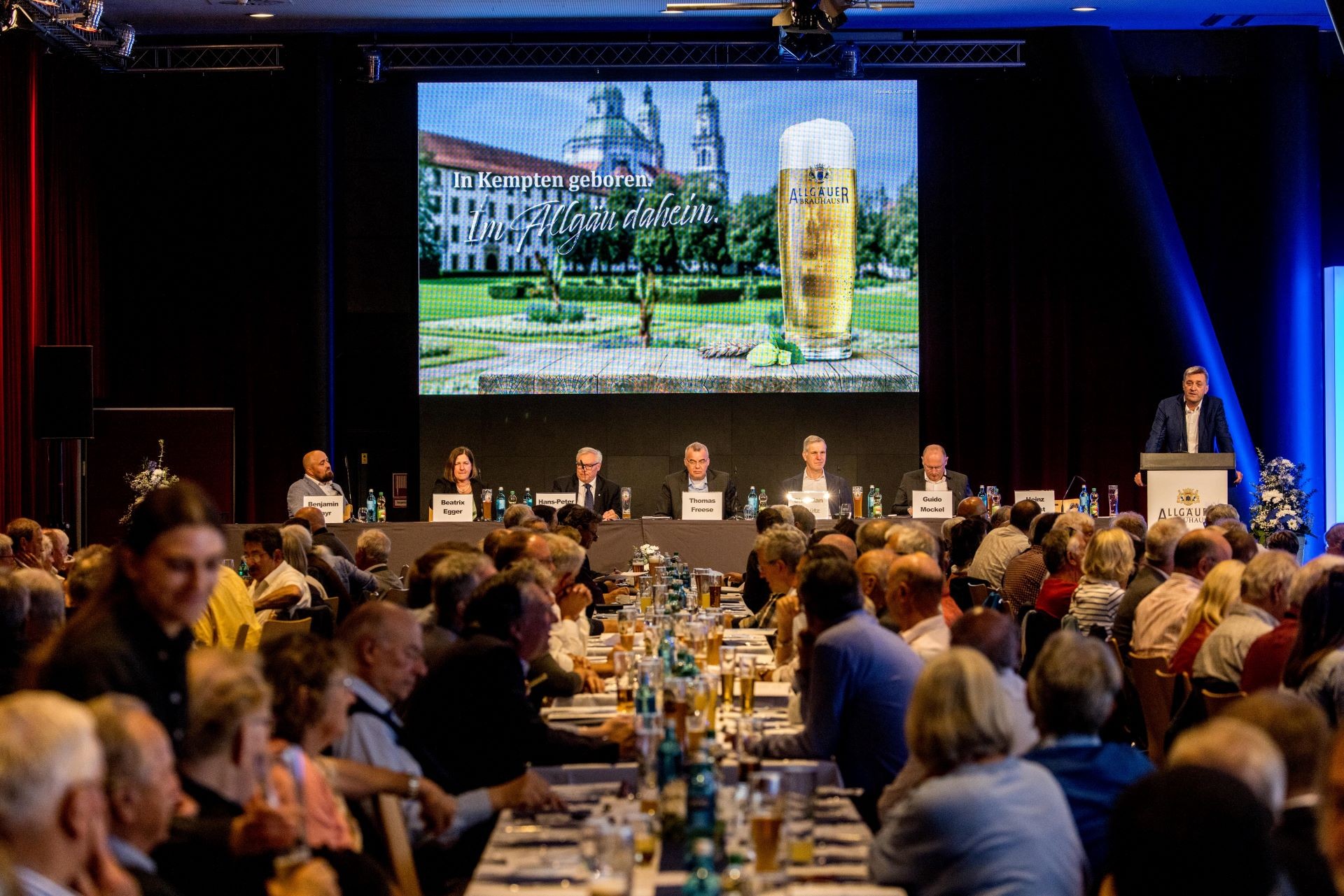 Mehrere Personen sitzen an Tischen während einer Konferenz, ein Mann spricht am Podium. Eine große Leinwand zeigt eine Werbeanzeige.