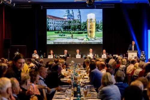 Mehrere Personen sitzen an Tischen während einer Konferenz, ein Mann spricht am Podium. Eine große Leinwand zeigt eine Werbeanzeige.