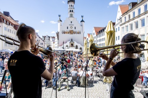 Zwei Personen spielen Posaune auf einer Bühne vor einer historischen Rathausfassade und einer Menschenmenge.