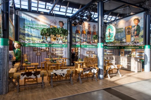 Innenansicht eines Messestands mit Themenbereich Alpen, einschließlich Holzdekoration und Bierzubehör.