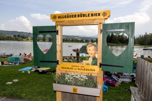 Holzschild mit Ausschnitt in Herzform vor einem belebten Seeufer mit Badegästen.