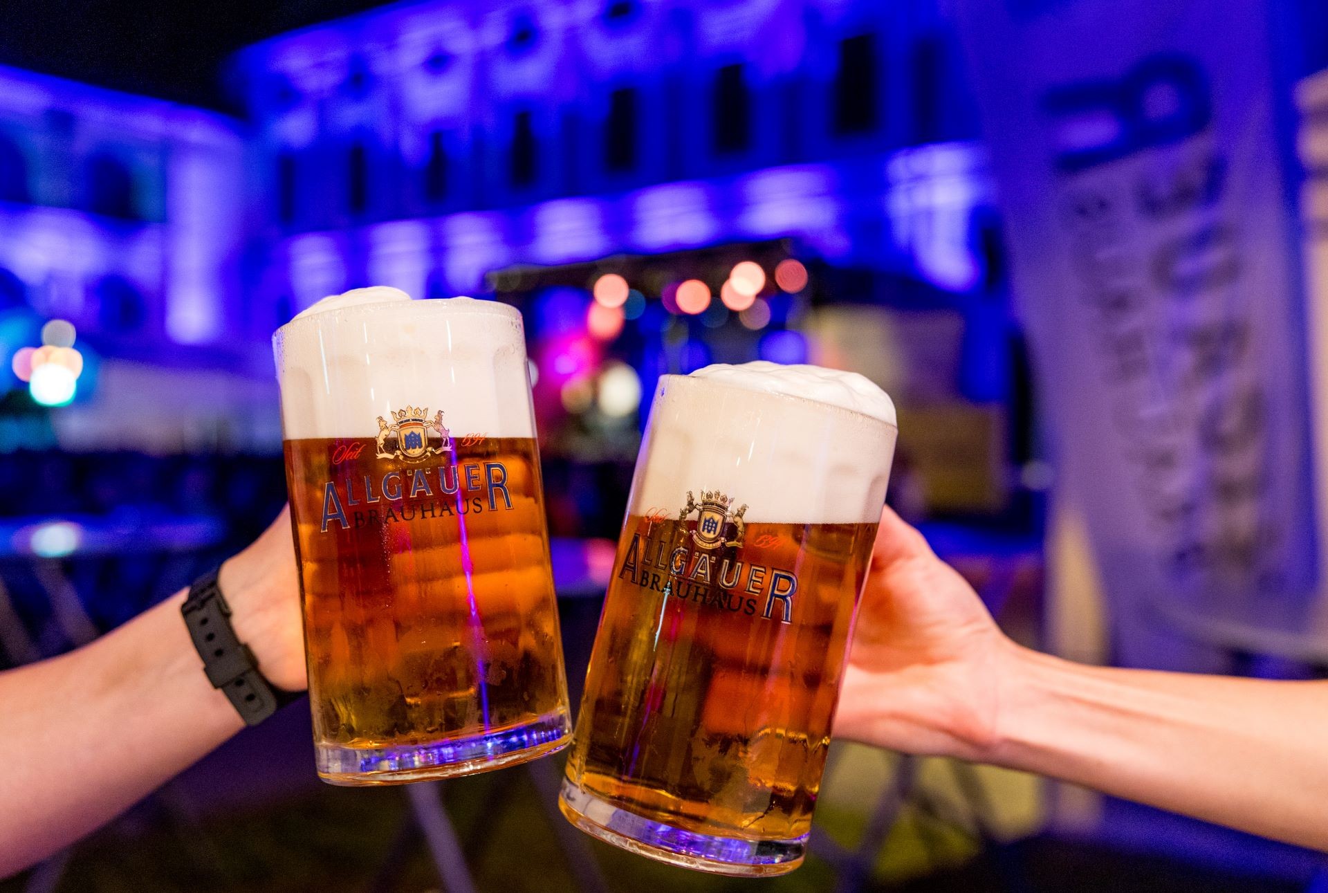Zwei Hände halten Biergläser vor einem beleuchteten Gebäude.