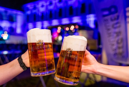 Zwei Hände halten Biergläser vor einem beleuchteten Gebäude.