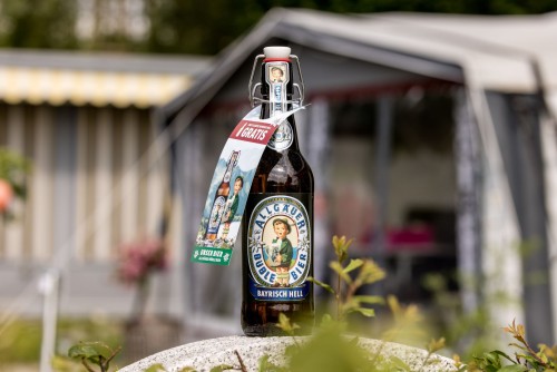 Eine verschlossene Bierflasche steht auf einem Stein im Freien. Sie trägt Etiketten mit traditionellen Motiven und Text.
