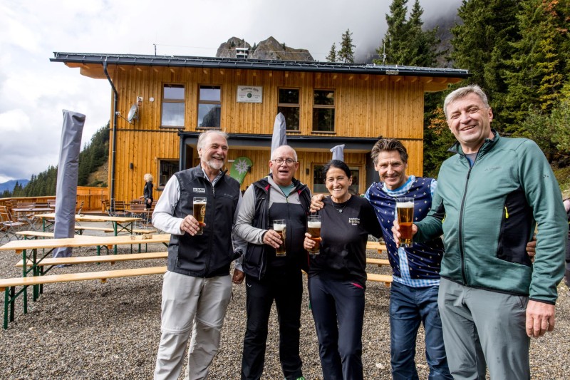 Fünf Personen lächeln mit Biergläsern vor einem Holzhaus im Gebirge.