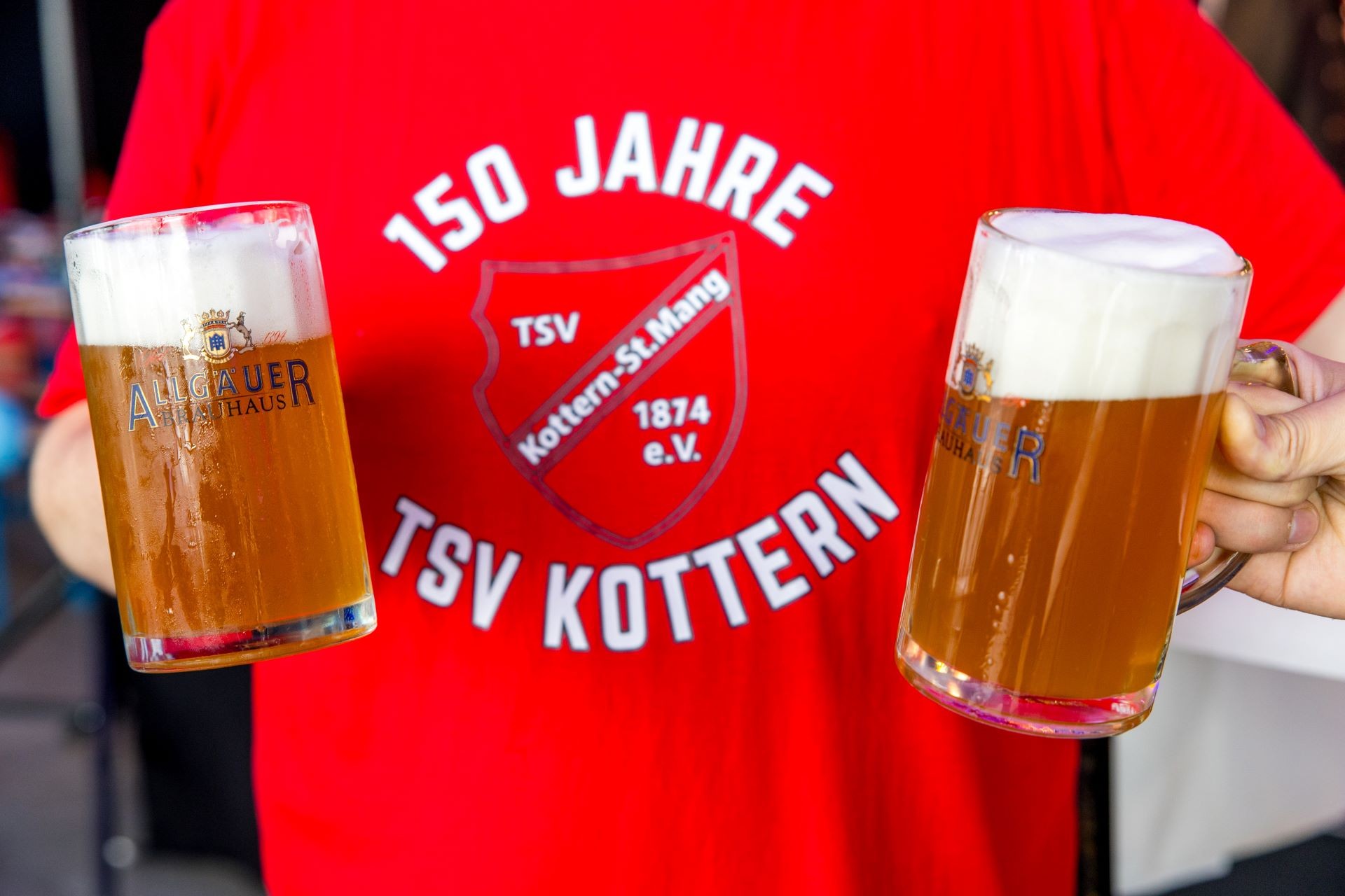 Eine Person in einem roten T-Shirt hält zwei Biergläser, gefüllt mit schaumigem Bier.