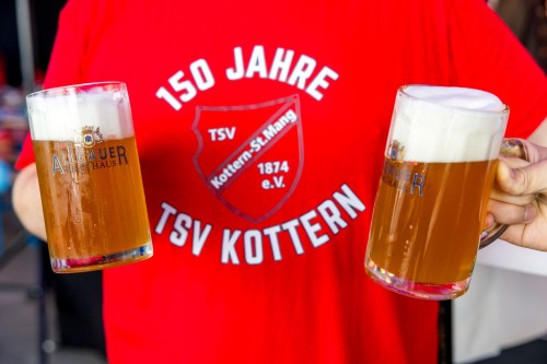 Eine Person in einem roten T-Shirt hält zwei Biergläser, gefüllt mit schaumigem Bier.