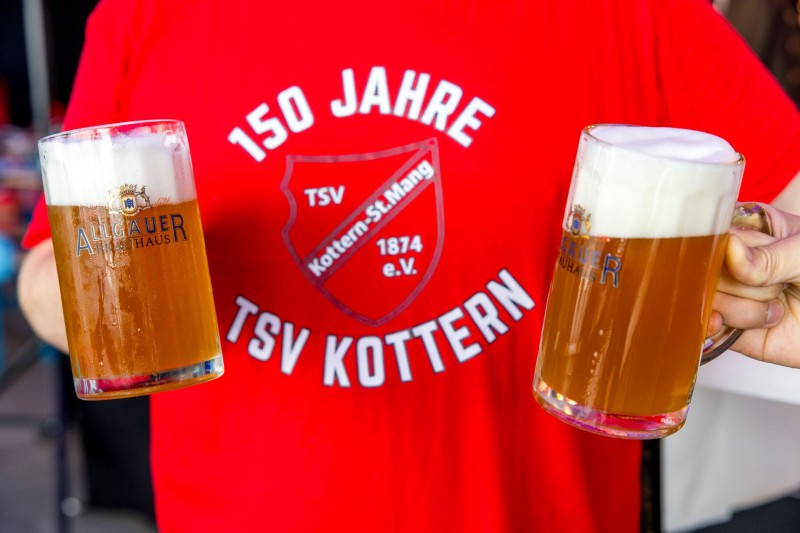 Eine Person in einem roten T-Shirt hält zwei Biergläser, gefüllt mit schaumigem Bier.