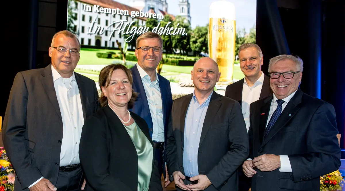 Sechs Personen in Businesskleidung stehen nebeneinander und lächeln in die Kamera. Im Hintergrund ein Allgäuer Brauhaus Motiv.