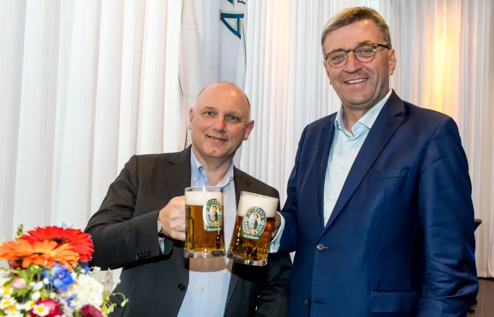 Zwei Männer im Anzug stoßen mit vollen Biergläsern an einem Stehtisch an.