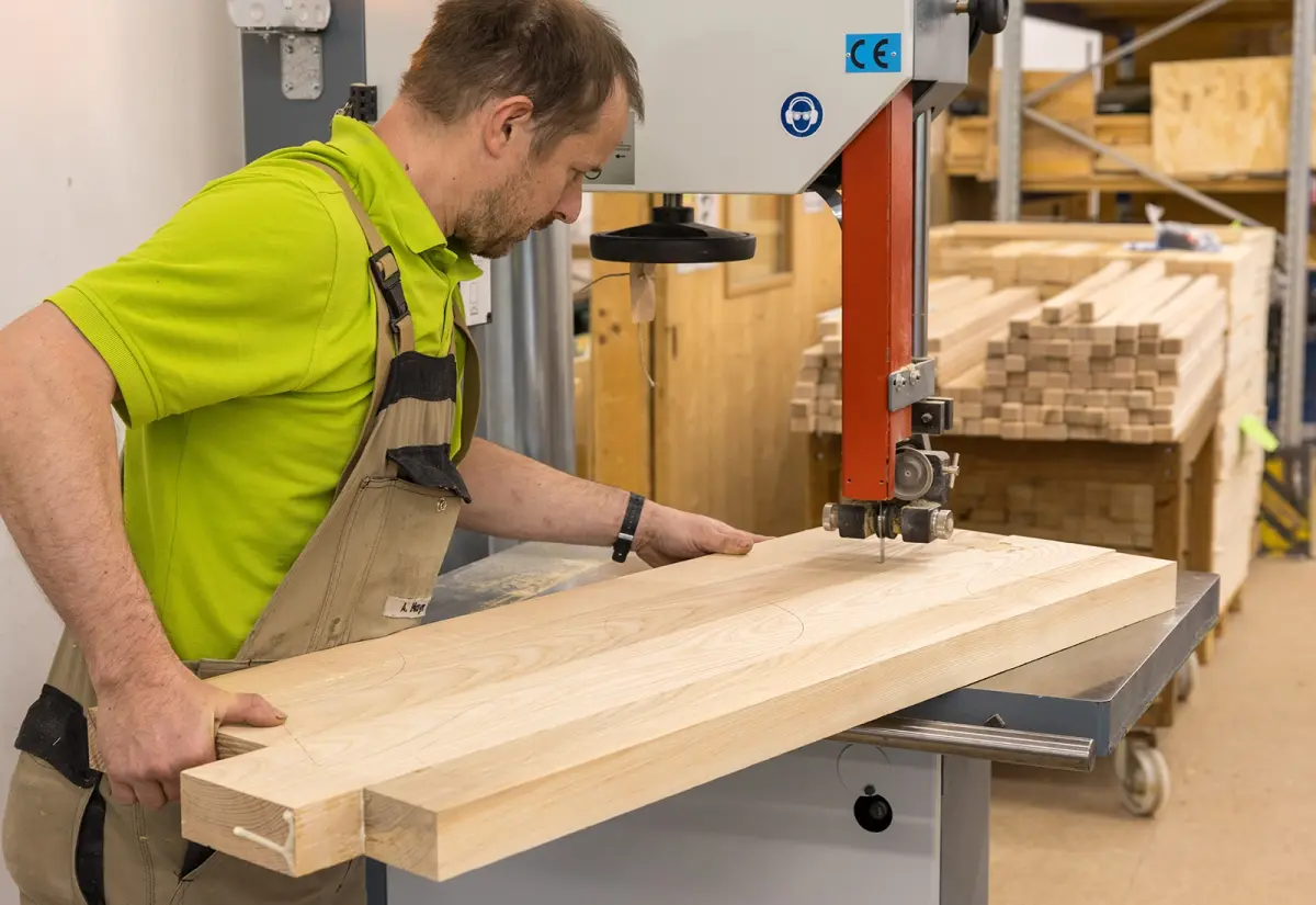 Ein Mann schneidet ein langes Holzbrett mit einer Bandsäge. Im Hintergrund liegen weitere zugeschnittene Holzteile.