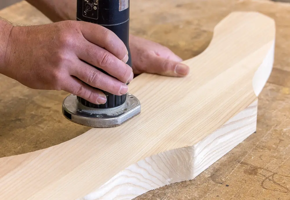Hände schleifen ein gebogenes Holzstück mit einer Maschine. Der Tisch besteht ebenfalls aus hellem Holz.