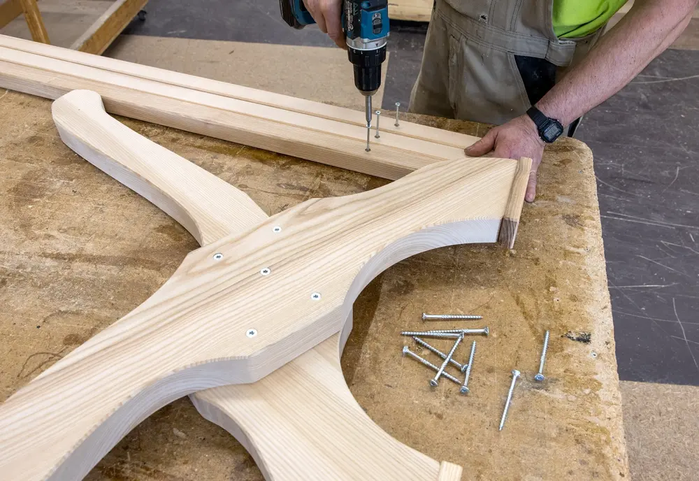 Ein Mann verschraubt zwei große Holzteile mit einer Bohrmaschine. Auf dem Tisch liegen weitere Schrauben.