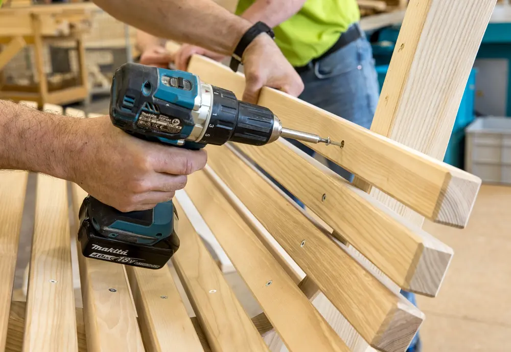 Zwei Personen montieren Holzlatten mit einer Bohrmaschine. Die Latten bilden eine Rückenlehne aus hellem Holz.