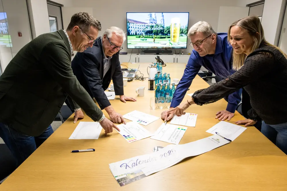 Vier Personen stehen an einem Konferenztisch und besprechen Auswertungsbögen. Im Hintergrund ist ein Bildschirm mit Biermotiv.