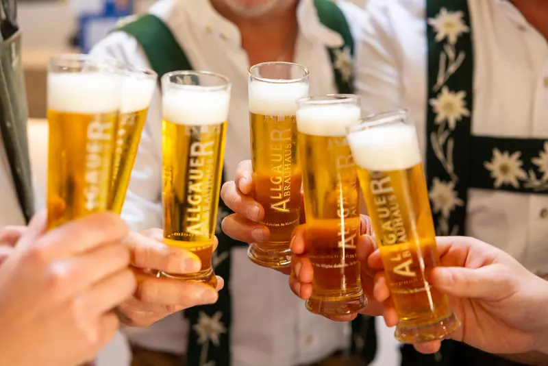 Gruppe in Trachten hält Allgäuer Brauhaus Biergläser zum Anstoßen hoch. Die Gläser sind mit Schaum bedeckt.