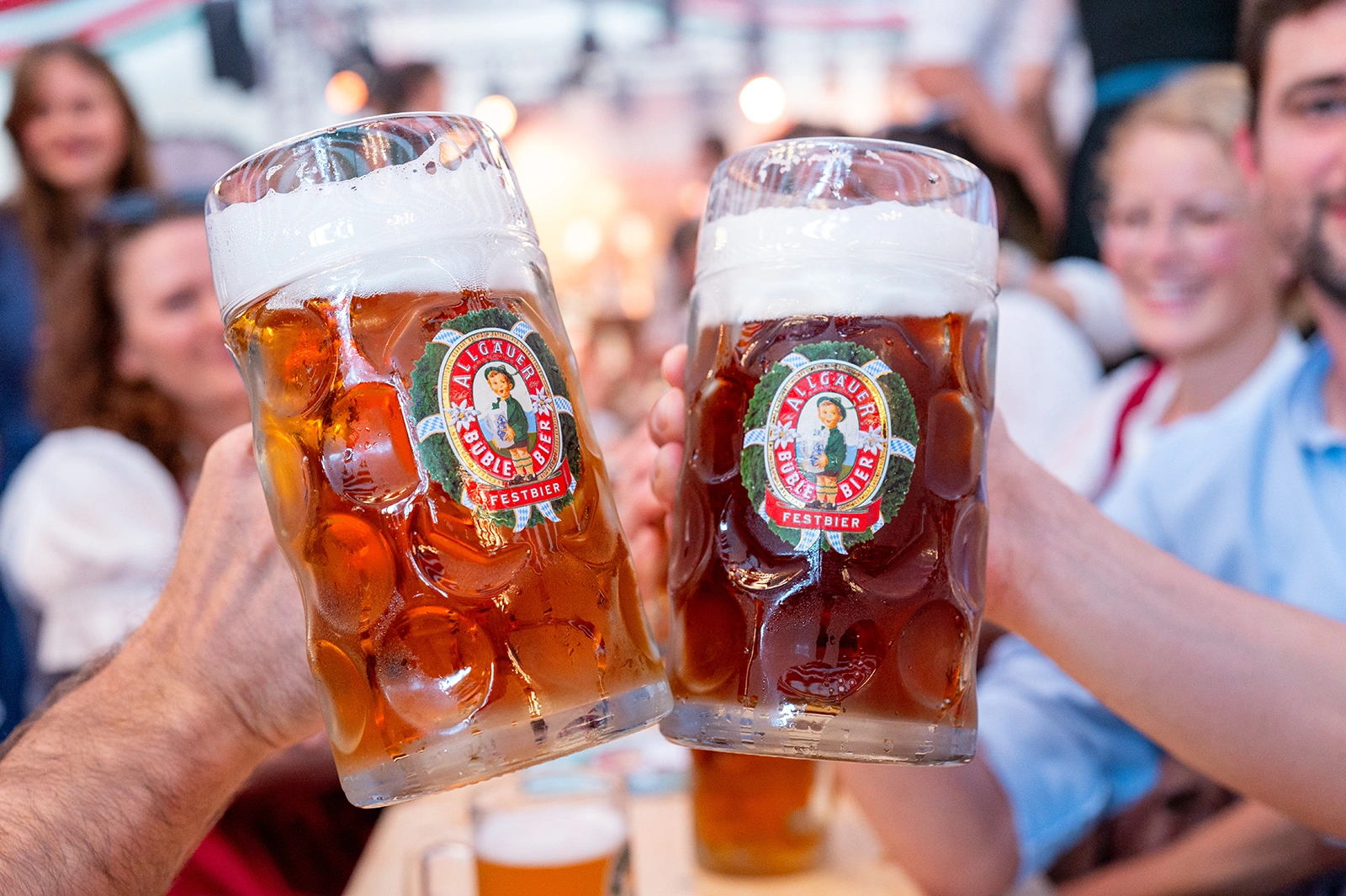 Große Allgäuer Büble Bier Bierkrüge mit Schaum werden von zwei Personen gehalten. Sie feiern in einer lebhaften Umgebung.