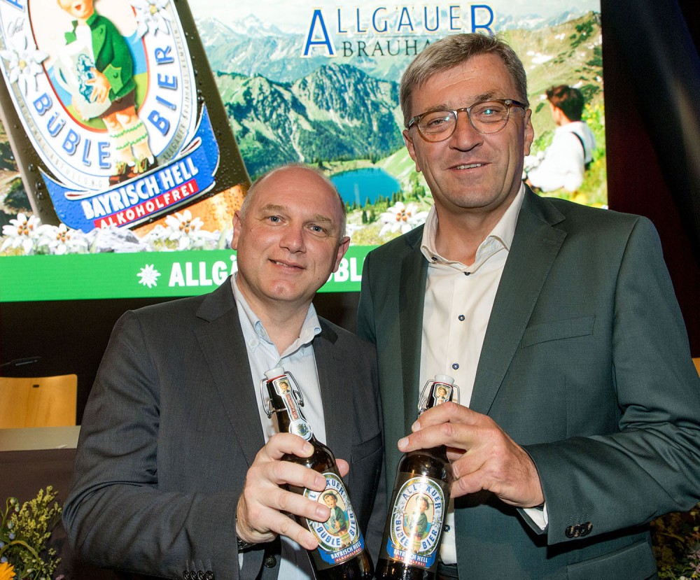 2 Männer stehen vor einer Büble Bier Bayrisch Hell Alkoholfrei Werbung und prosten mit 2 Bierflaschen.